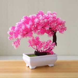 Un bonsai artificiale di fiori di ciliegio rosa brillante in un vaso bianco su un tavolo di legno chiaro davanti a una parete bianca.