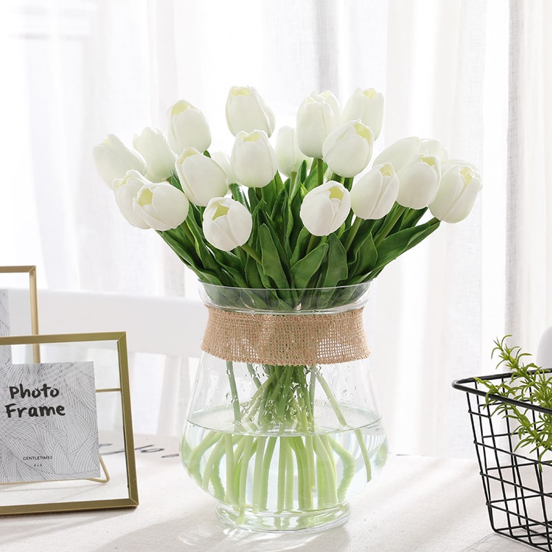 Un bouquet di tulipani bianchi in un vaso trasparente su un tavolo con una tovaglia bianca, una cornice e una tenda bianca alle spalle