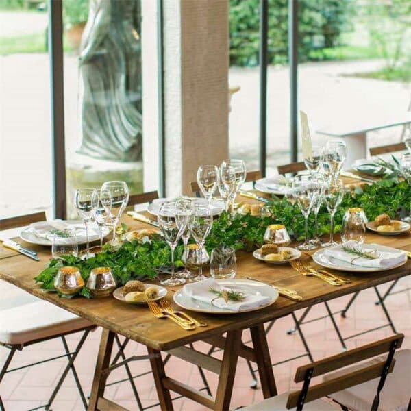 Tavolo in legno con coperti, candele dorate e una ghirlanda di foglie di edera sul runner del tavolo.
