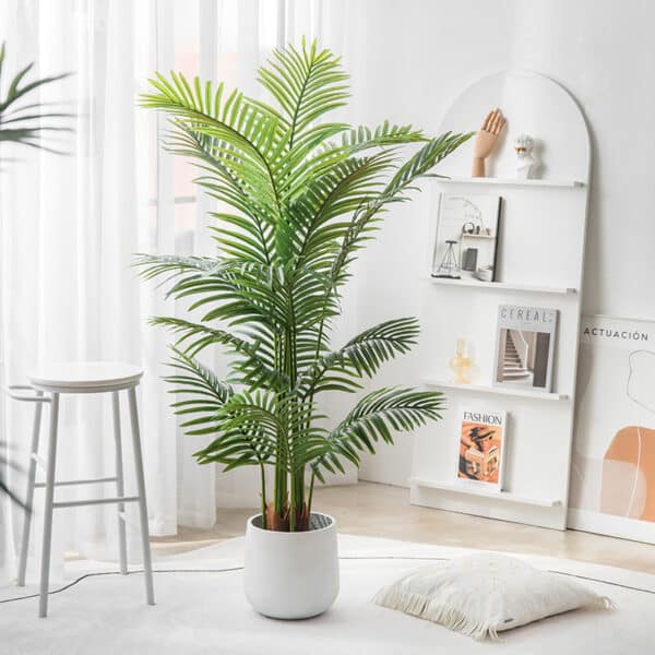 Una palma artificiale in un vaso bianco su un tappeto, accanto a un cuscino e a uno sgabello. Uno scaffale con 3 libri e tende