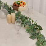 Un ramo di edera su un tavolo con una tovaglia bianca, bicchieri con stelo, candele e una torta di frutta.